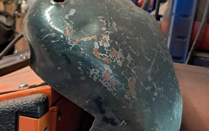 Old green motorcycle engine cover showing chipped paint and rust spots before restoration, placed on a workshop bench.