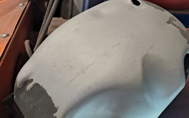Sandblasted motorcycle engine cover with smooth grey surface, resting on top of an orange blasting cabinet in a workshop.