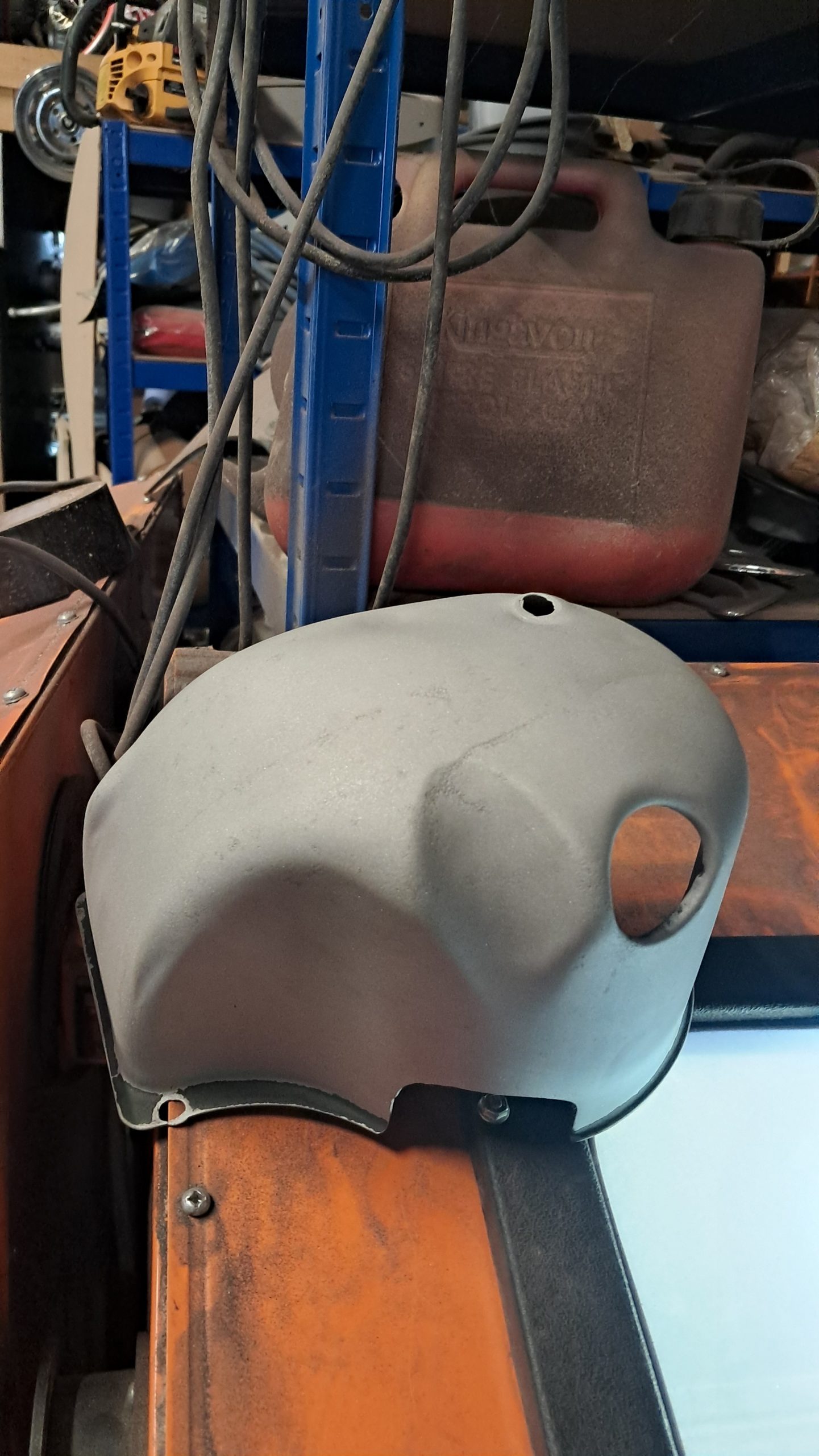 Grey sandblasted engine cover with circular and bolt holes, cleaned and ready for priming in a workshop setting.