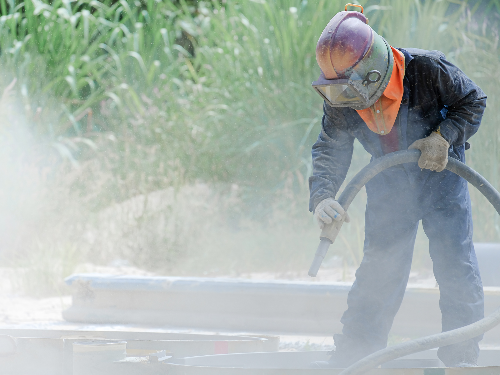 Worker wearing full protective gear and helmet operating a vapour blasting hose outdoors, surrounded by mist and dust, with greenery in the background.