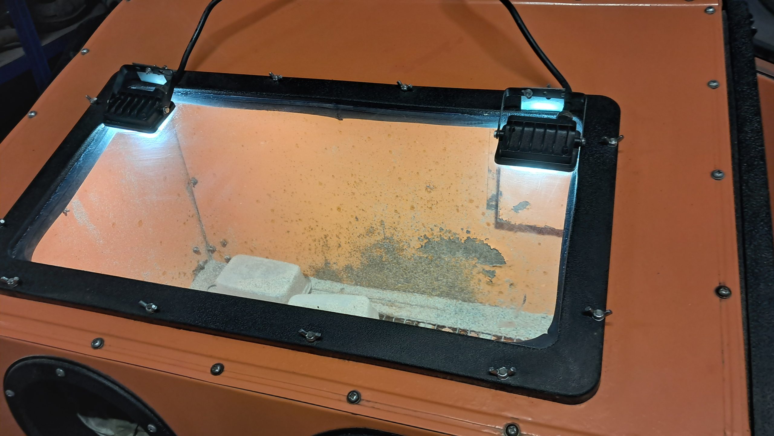 Side view of an orange blasting cabinet in a workshop surrounded by restoration tools and equipment.
